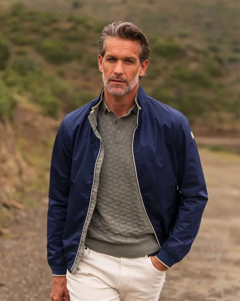 Man in a blue jacket and gray shirt stands outdoors on a gravel path, with green hills in the background.