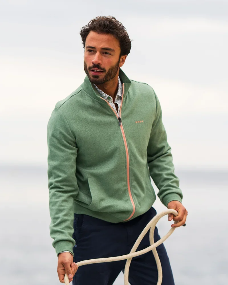Man with beard in a green zip-up jacket and blue pants holds a rope, looking to the side against an outdoor backdrop.